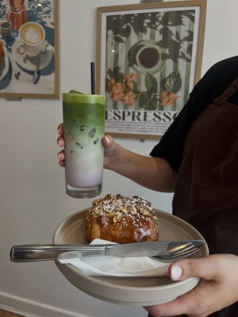 An iced matcha latte in a Lara Patisserie cup held up beside a cinnamon pastry